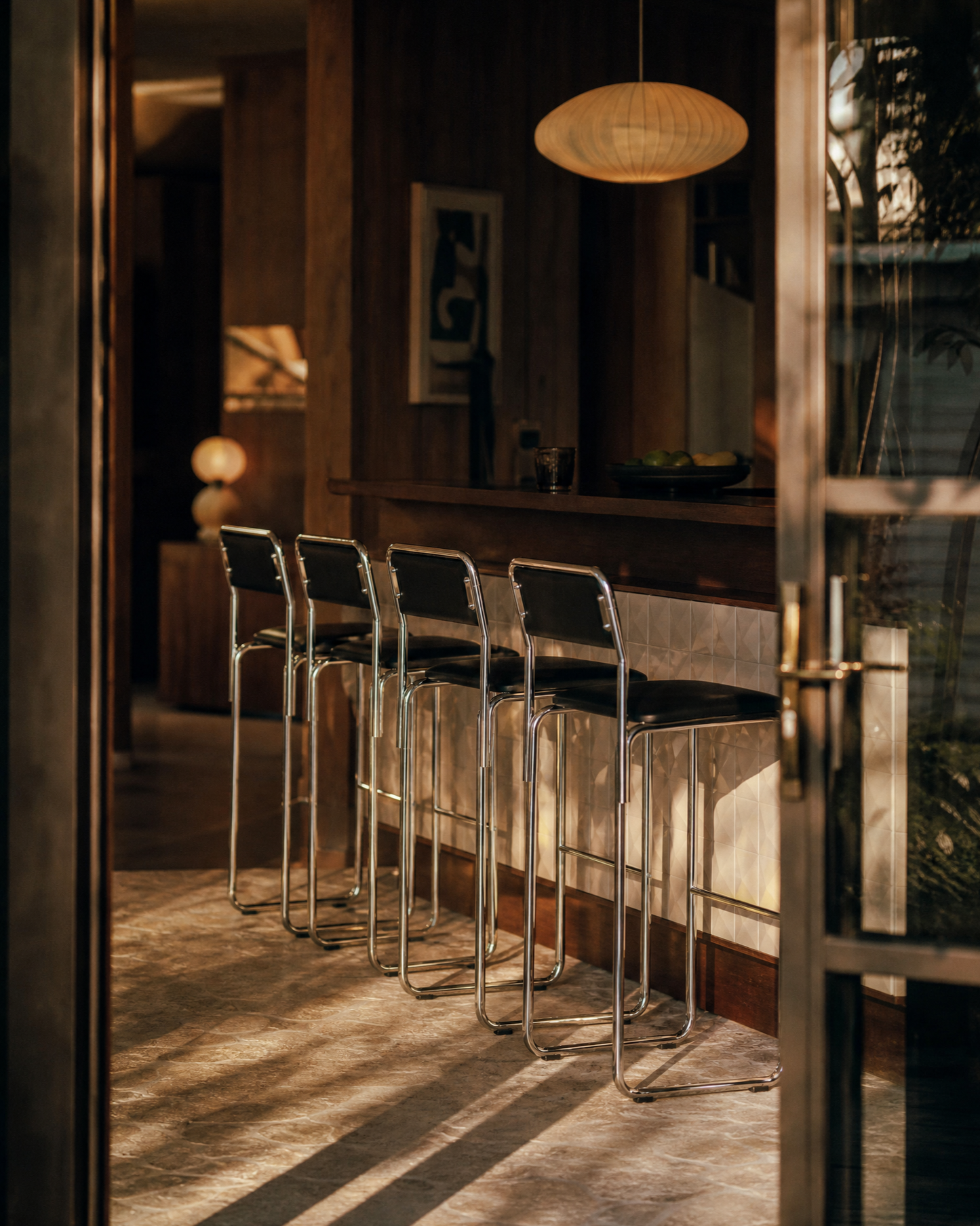 Four modern bar stools with chrome metal frames and black seats lined up at a wooden counter in a warmly lit interior space.