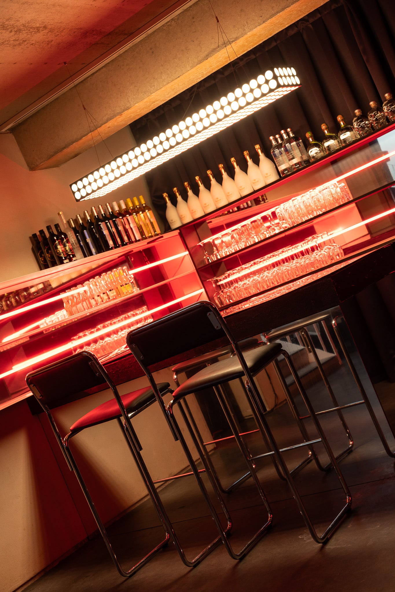 A modern bar interior with a long counter, illuminated red neon lighting, shelves of bottles and glasses, and several black bar stools in the foreground.