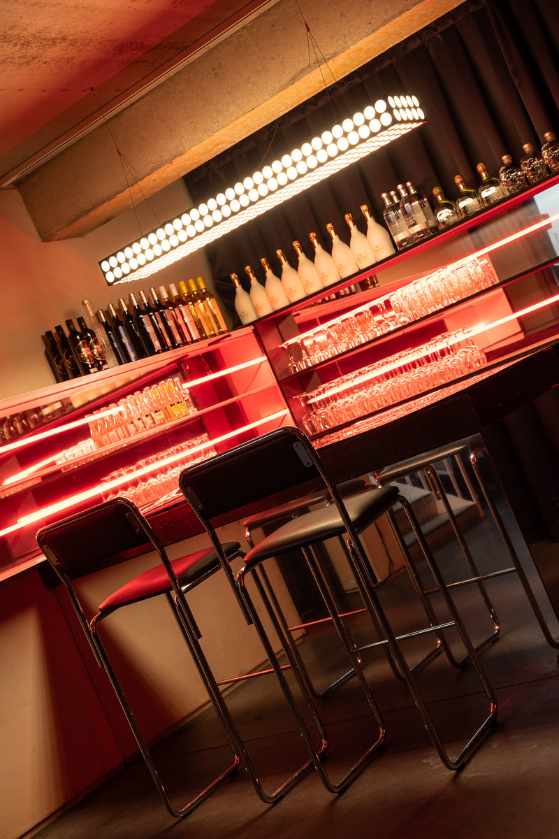 A modern bar interior with a long counter, illuminated red neon lighting, shelves of bottles and glasses, and several black bar stools in the foreground.